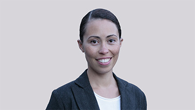 Nieves Martinez Marshall, a person with short dark hair slicked back wearing a dark gray blazer over a white top, against a gray background.